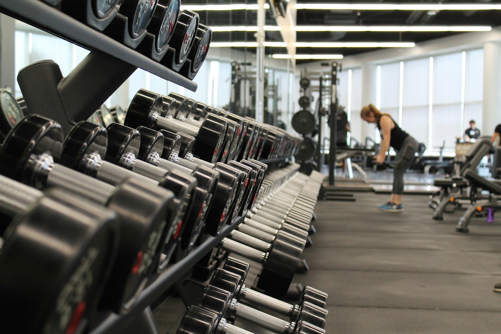Rack of dumbbells on a gym floor with a member training in the background